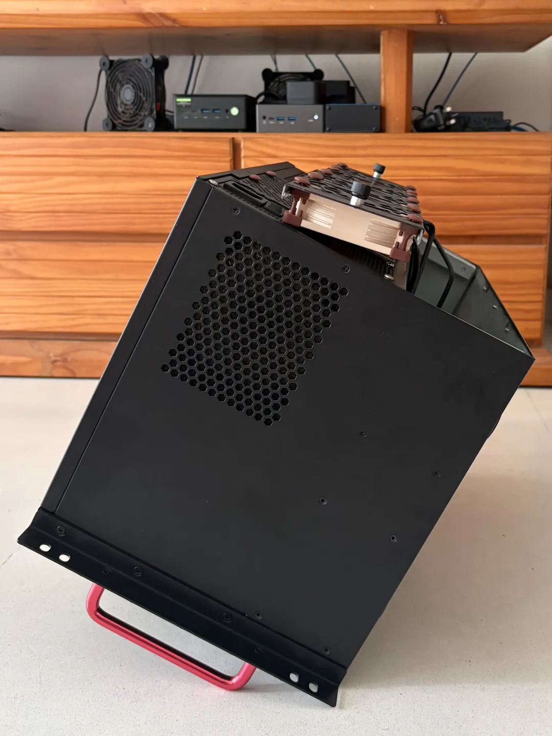 Black computer server rack case with back exposed with honeycomb vent, tilted on a red handle, against wooden cabinet.